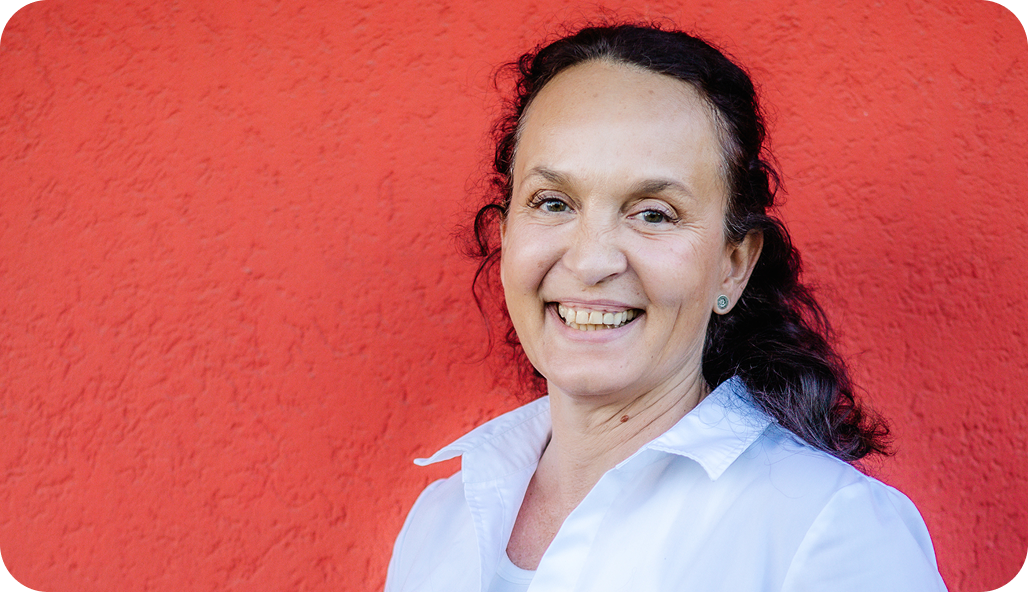 Dr. med. Karin Forschner steht lächelnd vor einer roten Wand mit schwarzen Haaren und einem weißen Hemd.
