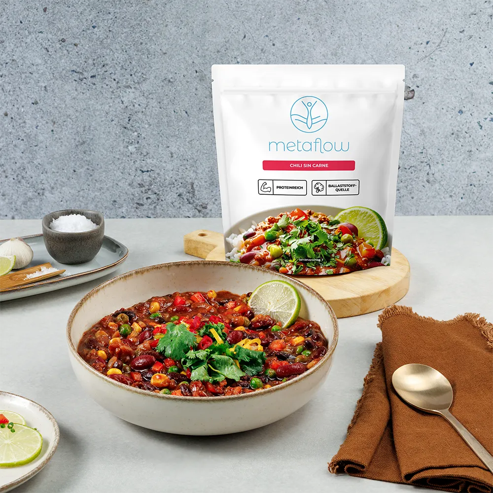 Eine Schüssel mit metaflow Chili sin Carne, gefüllt mit bunten Bohnen, Mais, Erbsen, Paprika und frischen Kräutern, garniert mit Limettenscheibe. Im Hintergrund steht die Produktverpackung mit dem metaflow-Logo und den Hinweisen ‚proteinreich‘ und ‚Ballaststoffquelle‘ auf einer hellen Tischoberfläche.