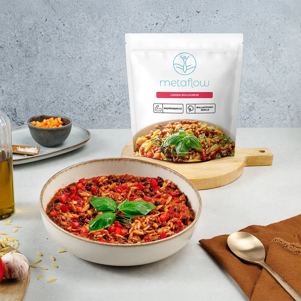Eine Schüssel mit metaflow Linsen Bolognese, angerichtet mit Linsen, Tomatensauce, Orzo-Nudeln und frischem Basilikum. Im Hintergrund steht die Produktverpackung mit dem blauen metaflow-Logo sowie den Kennzeichnungen ‚Proteinquelle‘ und ‚Ballaststoffquelle‘, auf einem hellen Küchentisch mit rustikalen Accessoires.