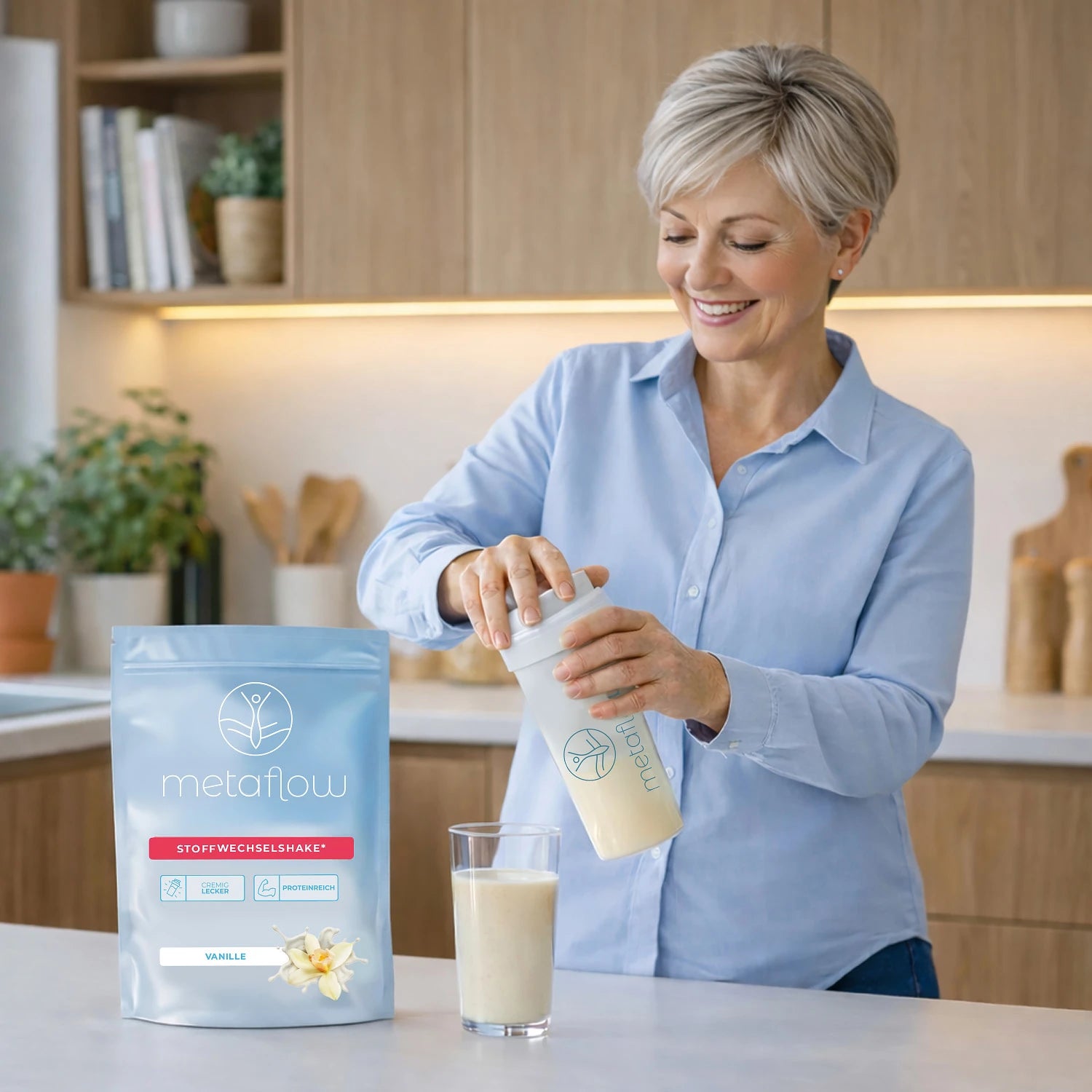 Eine lächelnde Frau mit kurzen grauen Haaren steht in der Küche und hat einen Shaker in der Hand. Vor ihr auf der Arbeitsfläche steht ein Beutel MetaFlow und ein Glas mit dem Stoffwechselshake.