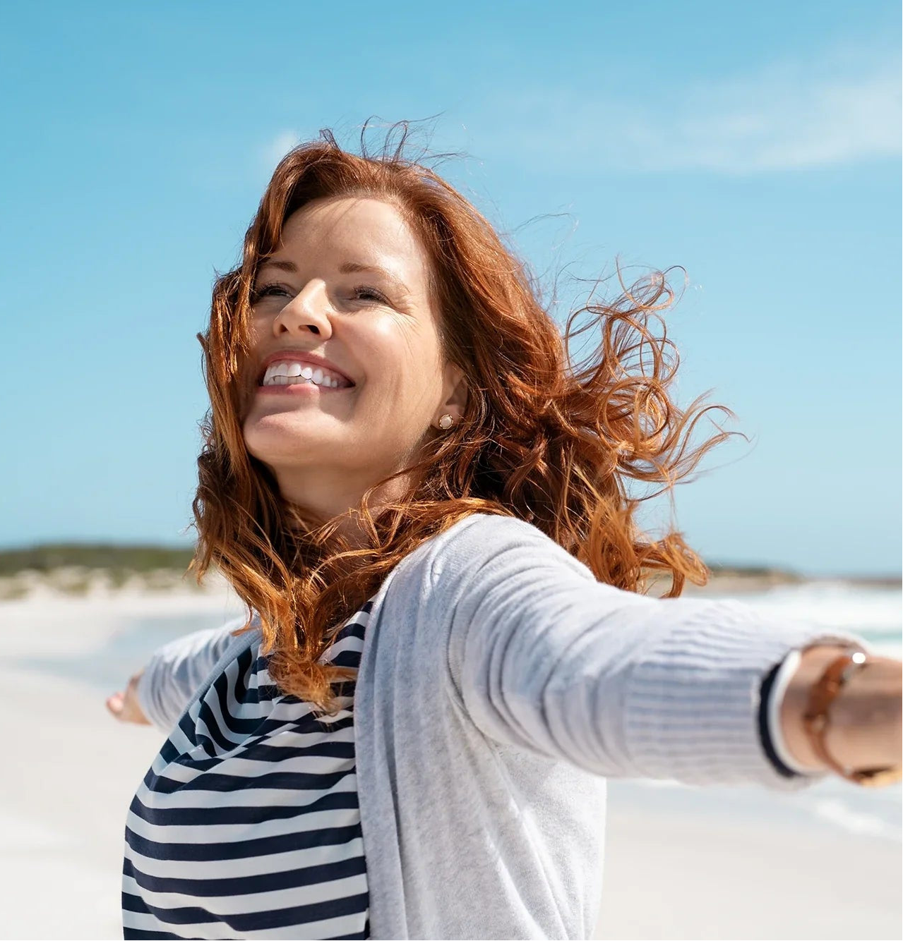 Lachende Frau mit roten Haaren und ausgebreiteten Armen, die am Strand in der Sonne steht, die Haare wehen im Wind