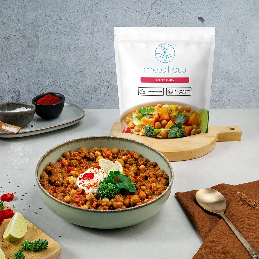 Eine Schüssel mit metaflow Gelbes Curry, bestehend aus Kichererbsen, Karotten, Brokkoli und Paprika, serviert mit Limettenscheibe, frischen Kräutern und Joghurt-Topping. Dahinter steht die passende metaflow-Verpackung mit den Symbolen ‚proteinreich‘ und ‚Ballaststoffquelle‘ auf einem Holzbrett.